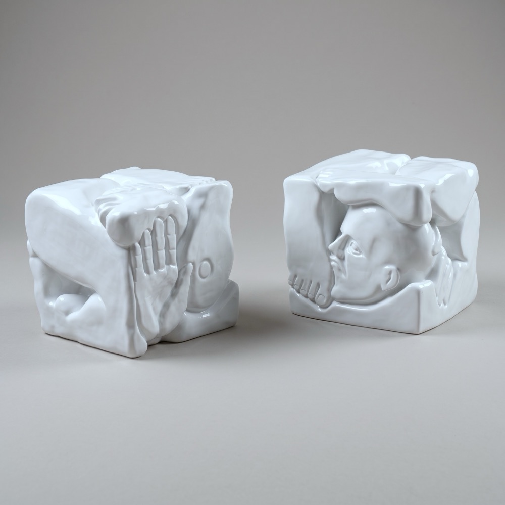 Two glossy white polyester cubes on the floor. Folded limbs and an open hand press against the sides of one; a bald head in profile, a shoulder and an arm wrap around the edges of the other.