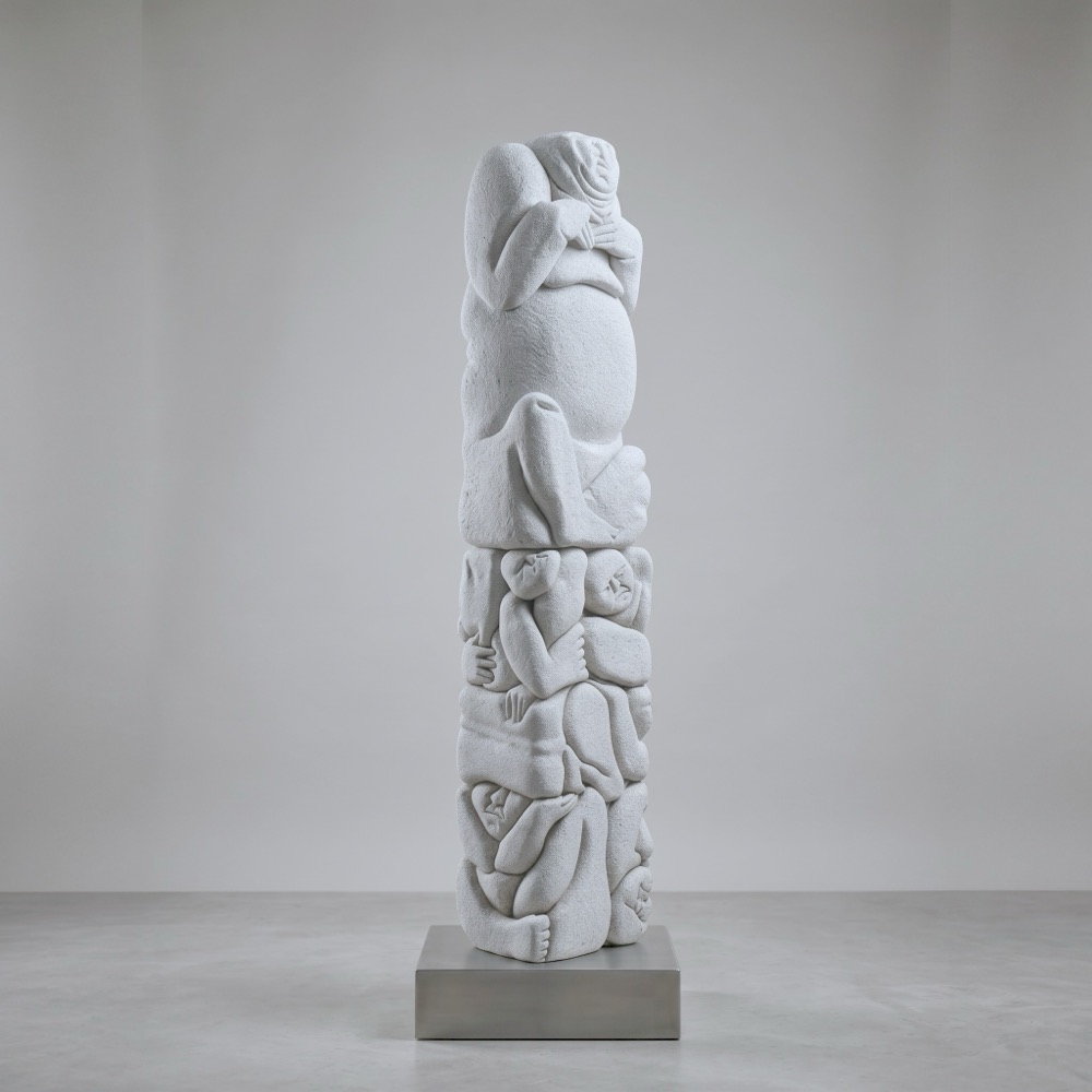 A tall, slender aerated-concrete column. A figure with a rounded belly sits at the top, carried by a dense mass of smaller interlocking bodies and faces below.